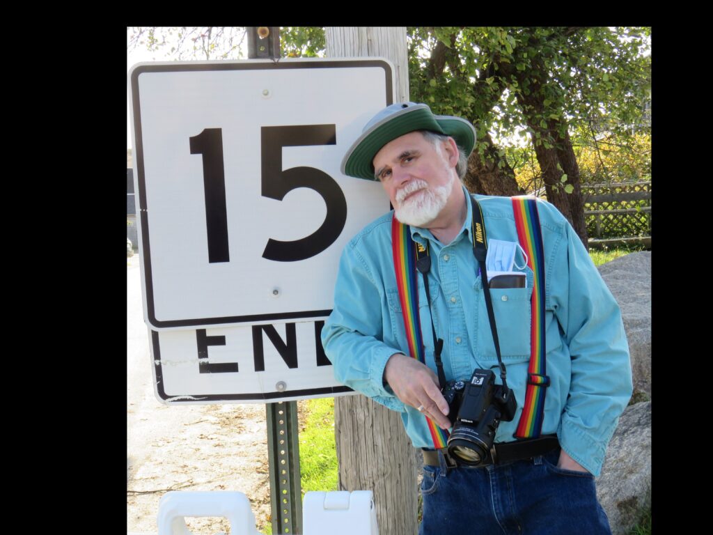 Picture of a man in a green denim shirt, with rainbow suspenders, camera around his neck wearing a hat, and leaning on a large route sign that says "15 END"